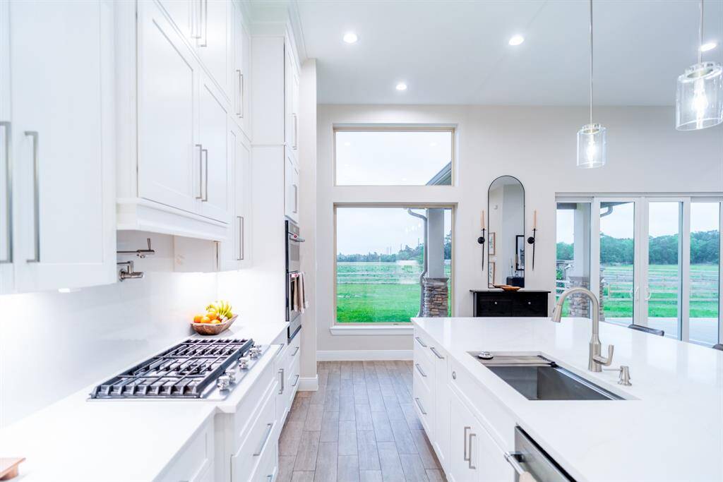 This modern kitchen features sleek white cabinetry, a gas stove, and a large island with a sink. It boasts ample natural light from expansive windows, providing a beautiful view of the outdoors.