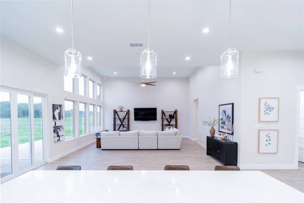 This spacious living area features an open floor plan with high ceilings and abundant natural light from large windows. It includes a modern design with a comfortable seating area, wall-mounted TV, and stylish lighting fixtures. The space opens to a scenic outdoor view, creating a seamless indoor-outdoor connection.