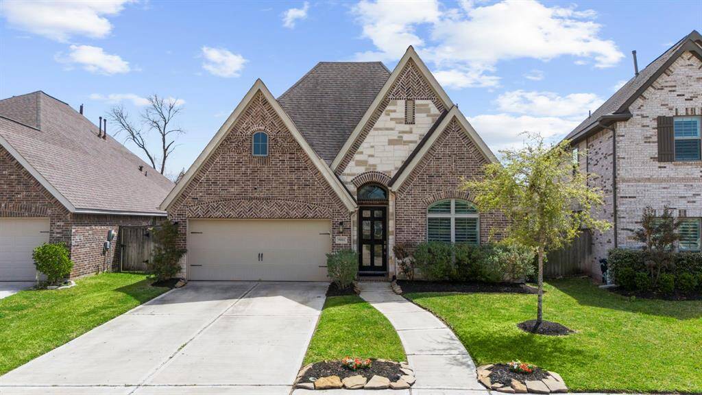 A charming brick home with a steep gable roofline, two-car garage, and a well-maintained front yard. The stone-bordered garden beds and curved walkway add curb appeal, along with symmetrical landscaping and a black front door with glass panels.