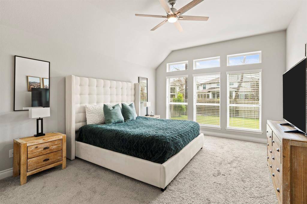 A spacious and serene primary bedroom with a plush upholstered bed, cozy carpet, and a large wall of windows that brings in natural light and views of the backyard. The neutral tones and pops of color from the bedding add a calm and modern touch.
