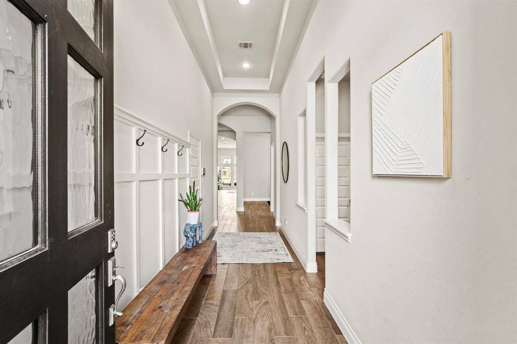A long, elegant hallway with wood-look tile flooring, wall paneling with hooks, and a rustic wooden bench. The space leads toward an open living area with arched doorways and recessed lighting.