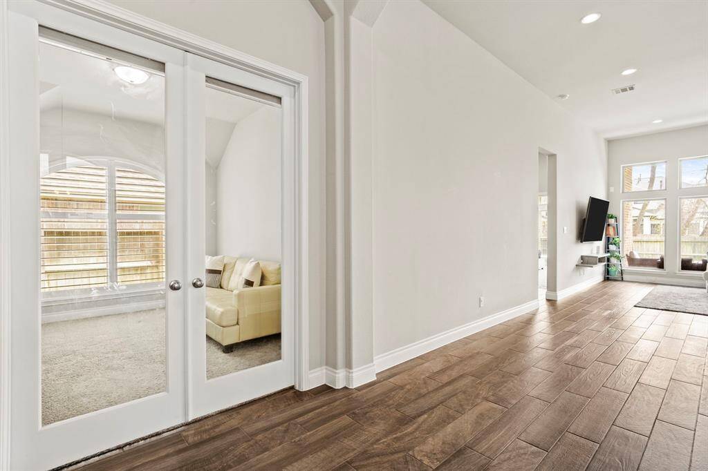 Glass-paned French doors open into a versatile space, currently staged as a sitting area or home office. The room features plush carpet, a large arched window, and clean neutral finishes.