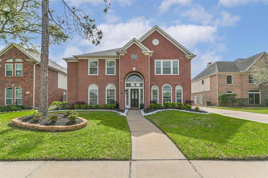 This is a two-story, red-brick home with a classic, symmetrical facade and arched windows. The well-maintained front yard features a curved walkway and landscaped flower beds, providing great curb appeal.