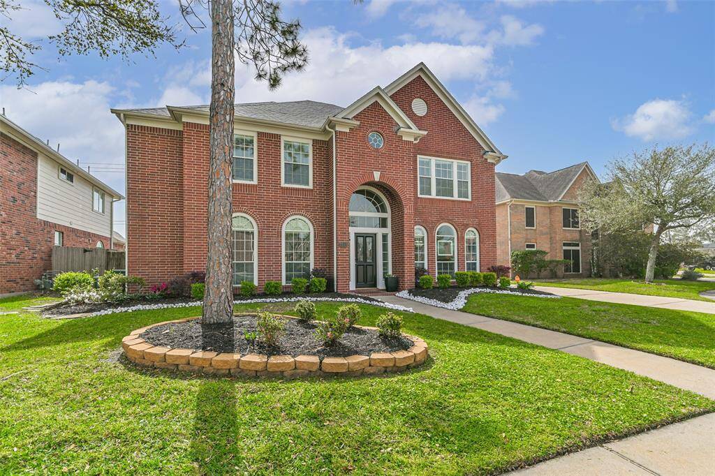 This is a two-story red brick home with a classic design, featuring large arched windows and a welcoming front entrance. It has a well-maintained lawn, landscaped garden beds, and a tree in the front yard, offering great curb appeal in a suburban neighborhood.