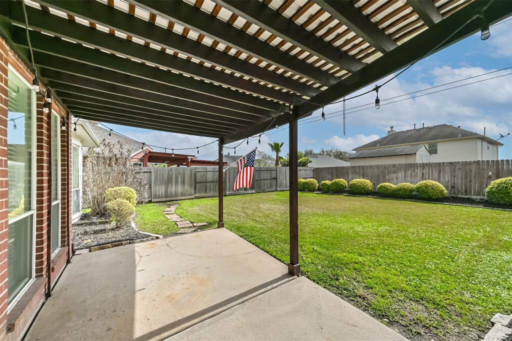 This backyard features a spacious covered patio with a pergola, perfect for outdoor gatherings. The well-maintained lawn is surrounded by a wooden fence, offering privacy and a charming garden space with shrubs. Ideal for relaxing or entertaining.