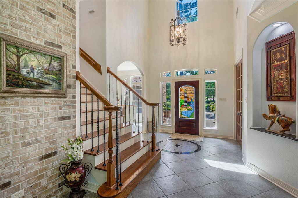 This elegant entryway features a grand staircase with wooden steps and iron railings, a high ceiling with a chandelier, and a vibrant stained glass front door. The space is accented with a brick wall, artwork, and decorative elements, creating a warm and inviting atmosphere.