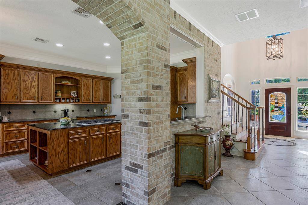 This photo showcases a spacious kitchen with warm wooden cabinetry and a large island, complemented by a brick archway. Adjacent is an elegant foyer featuring a staircase and a front door with decorative stained glass, creating a welcoming and stylish entryway.