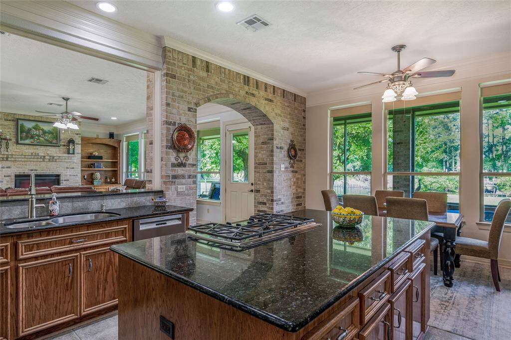 This cozy home features a spacious open-concept kitchen with a large island, granite countertops, and a gas cooktop. The kitchen flows into a bright dining area with ample natural light and views of the lush greenery outside. A brick archway leads to a comfortable living room with a fireplace, perfect for family gatherings.