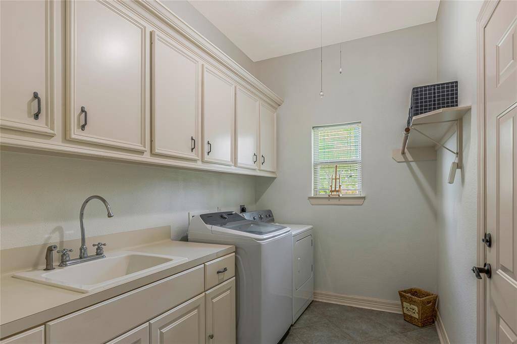 This is a spacious, well-lit laundry room featuring ample cabinetry, a sink, and a washer-dryer set. There's a small window for natural light and a shelf for extra storage.