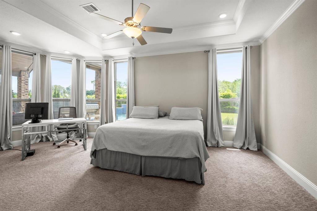 The spacious primary bedroom features a wall of windows overlooking the backyard, a cozy sitting area, and tiered ceilings with a ceiling fan and crown molding.