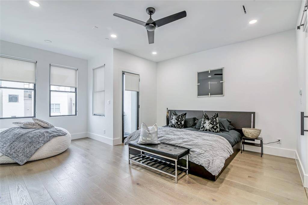 This modern bedroom features sleek hardwood floors, a ceiling fan, and ample natural light from multiple windows. The space includes a cozy bed setup with stylish decor and a comfortable seating area. Perfect for a contemporary and relaxed living environment.