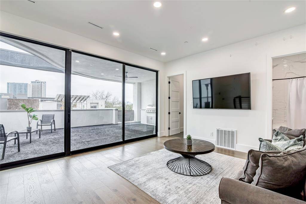 This modern living space features an open layout with large glass doors leading to a spacious balcony, providing abundant natural light and city views. It includes sleek hardwood floors, a wall-mounted TV, and a cozy seating area, perfect for relaxation and entertainment.