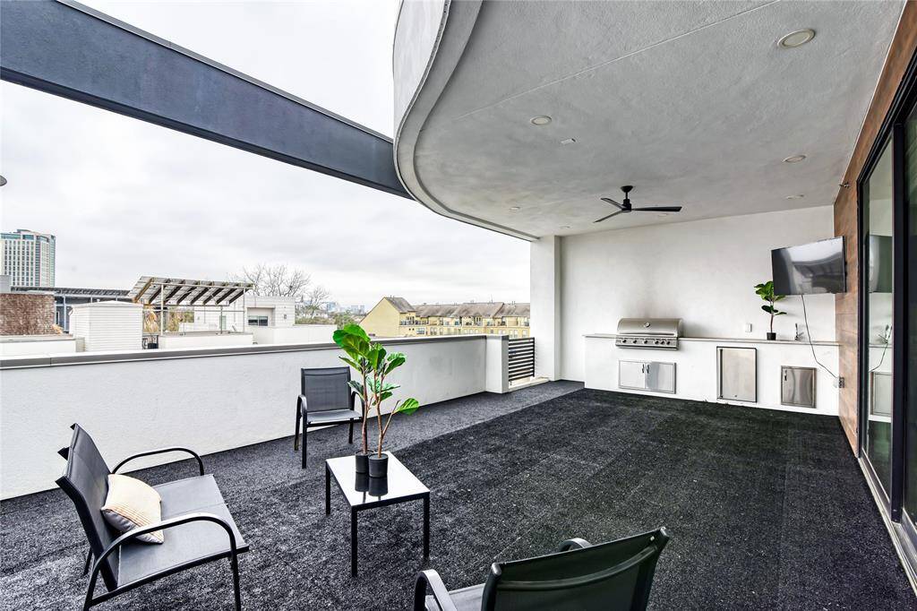 This photo showcases a spacious covered patio with modern outdoor furniture, a built-in grill, and a TV. It features a ceiling fan and has views of the surrounding neighborhood, perfect for entertaining or relaxing.