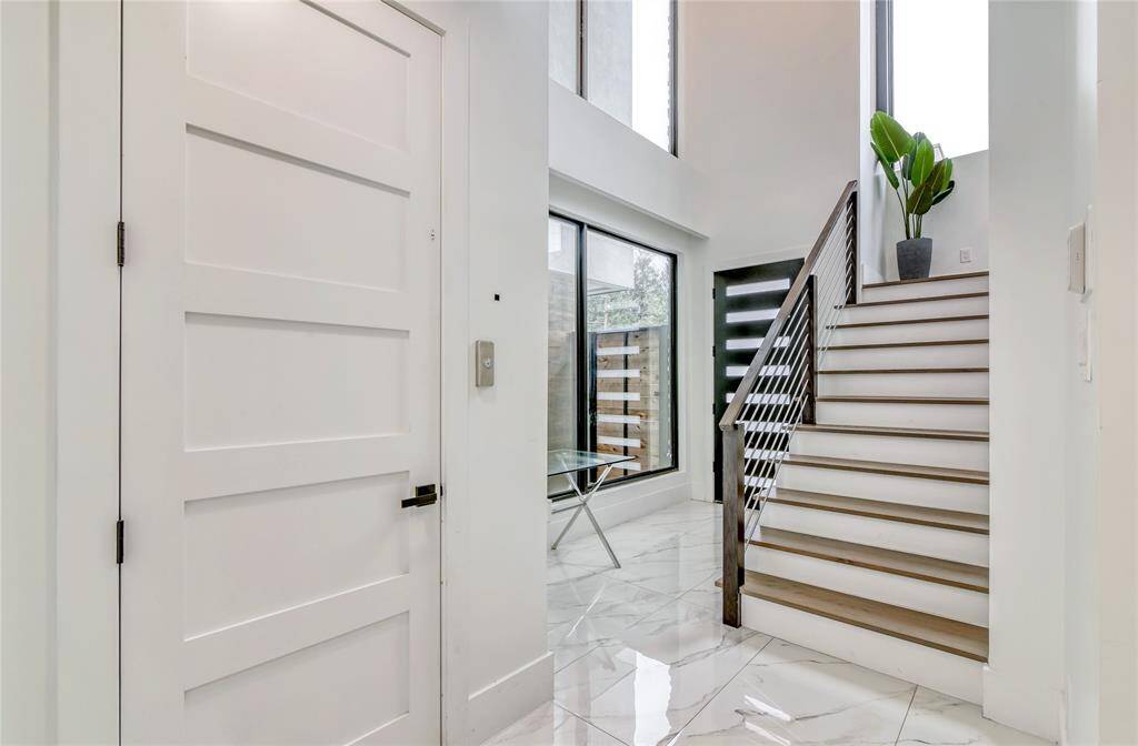 This home features a modern entryway with sleek, white paneled doors and glossy marble flooring. A stylish staircase with wooden steps and metal railing leads to the upper level, complemented by large windows that bring in natural light.
