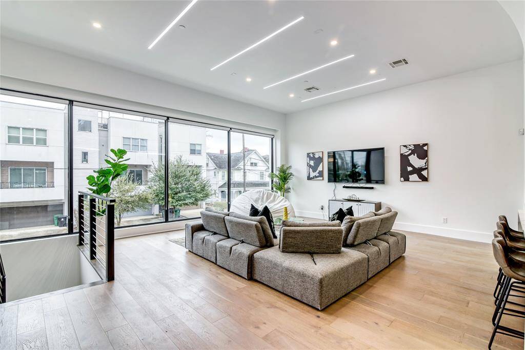 This modern living room features an open layout with sleek bamboo floors and abundant natural light from large windows. It includes a contemporary sectional sofa, wall-mounted TV, and stylish decor, creating a bright and inviting space.