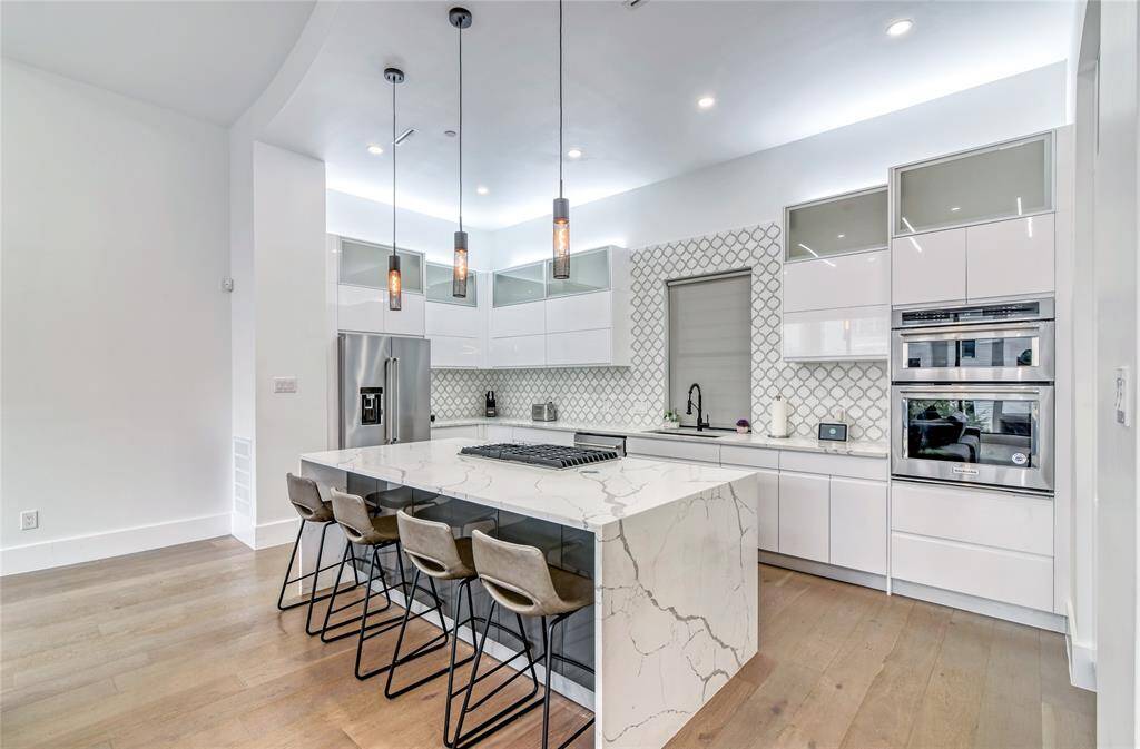 This modern kitchen features a sleek design with white cabinetry, a marble island with bar seating, and stainless steel appliances. The backsplash has a stylish geometric pattern, and pendant lighting adds a warm touch.