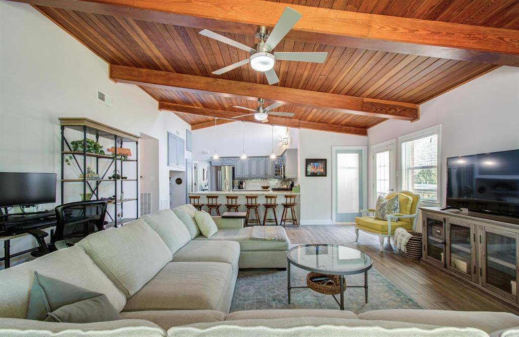 This spacious living area boasts a warm wooden ceiling with exposed beams, an open layout, and an abundance of natural light, creating a bright and airy atmosphere. The generous floor plan flows seamlessly into a stylish kitchen with an island and barstool seating, offering ample space for both relaxation and entertaining.