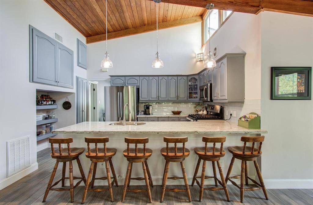 This kitchen features a spacious layout with high ceilings, a warm wooden ceiling accent and windows that bring in lots of natural light. It includes modern stainless steel appliances, gray cabinetry, and a large island with seating for six. Pendant lighting adds a stylish touch, and open shelving offers additional storage.