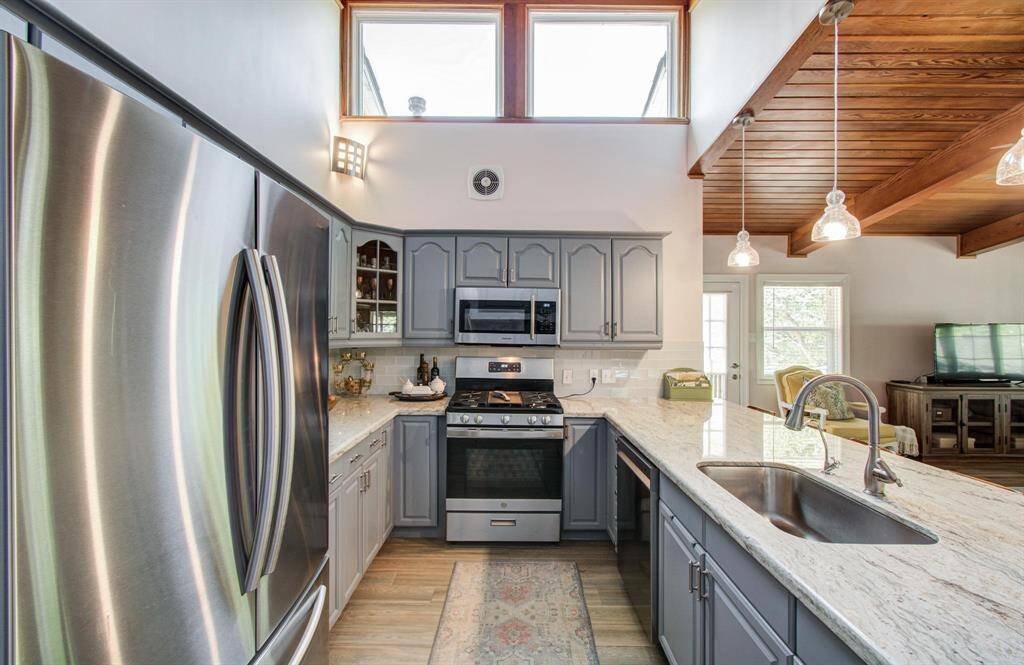 Modern kitchen with stainless steel appliances, granite countertops, and stylish gray cabinets. High ceilings and large windows provide ample natural light. Open layout with wooden ceiling accents and pendant lighting adds warmth and character.