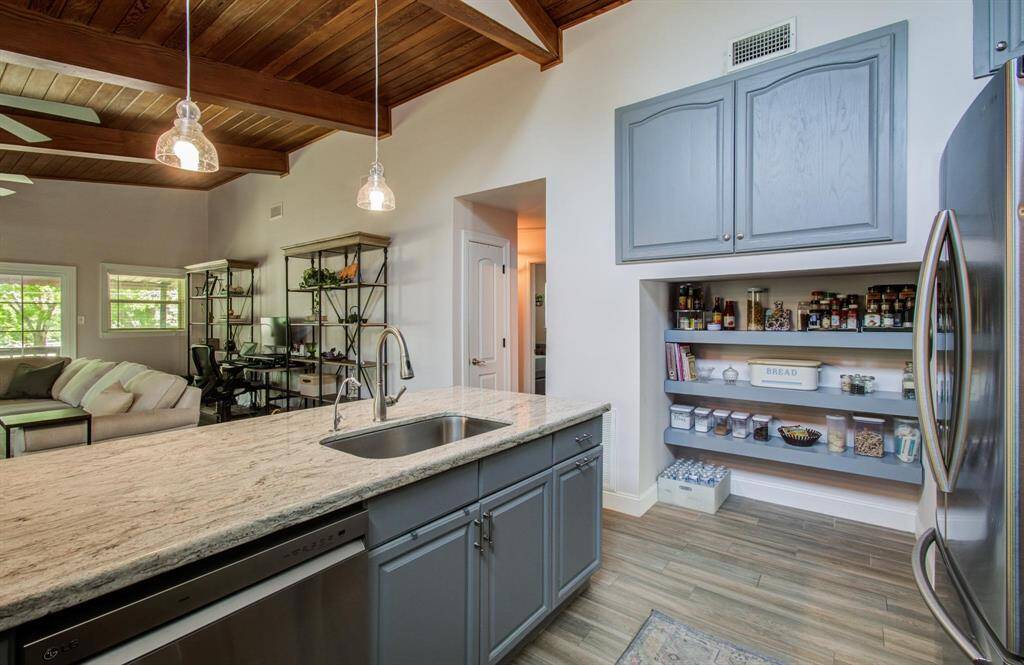 The modern kitchen features shaker-style gray cabinets and a marble countertop, blending rustic charm with sleek design. Open shelving offers practical storage, while stainless steel appliances and pendant lighting add a touch of contemporary style. The space flows seamlessly into a cozy living area with wood-beamed ceilings, creating a warm and inviting atmosphere with a perfect blend of modern and rustic elements.