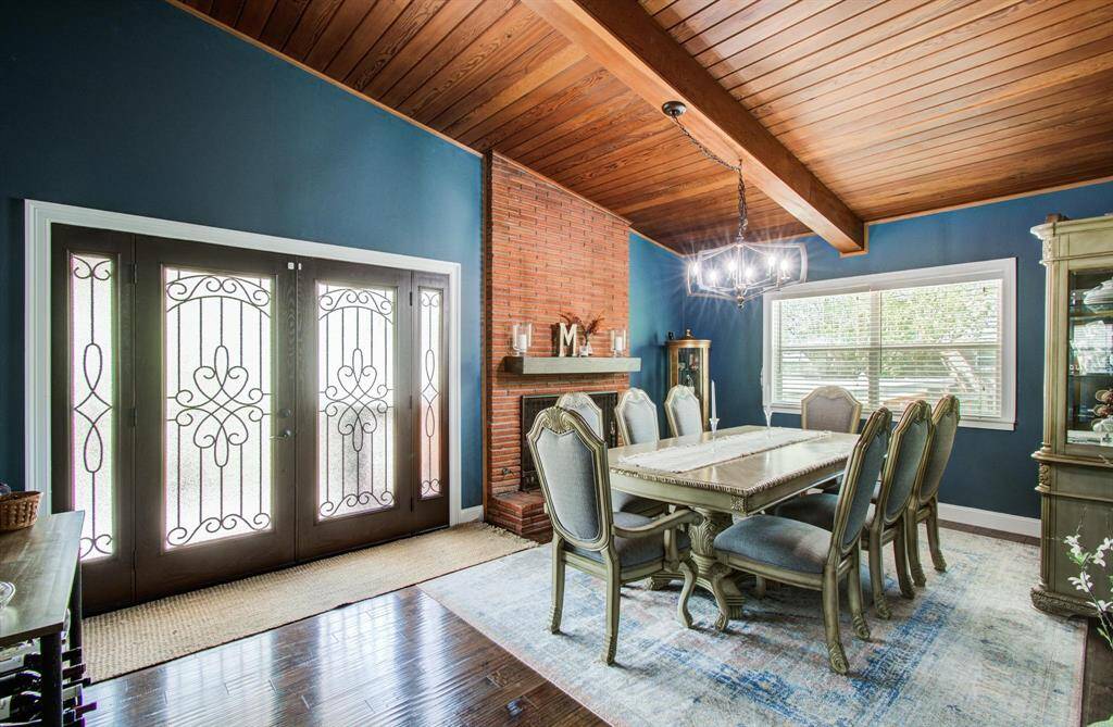 This cozy formal dining room exudes warmth and character, centered around a classic brick fireplace that invites intimate gatherings. The wooden ceiling with exposed beams adds depth and texture, while the rich blue walls create a sense of elegance and calm. Natural light pours in through a large window, softening the space and highlighting its inviting, timeless appeal.