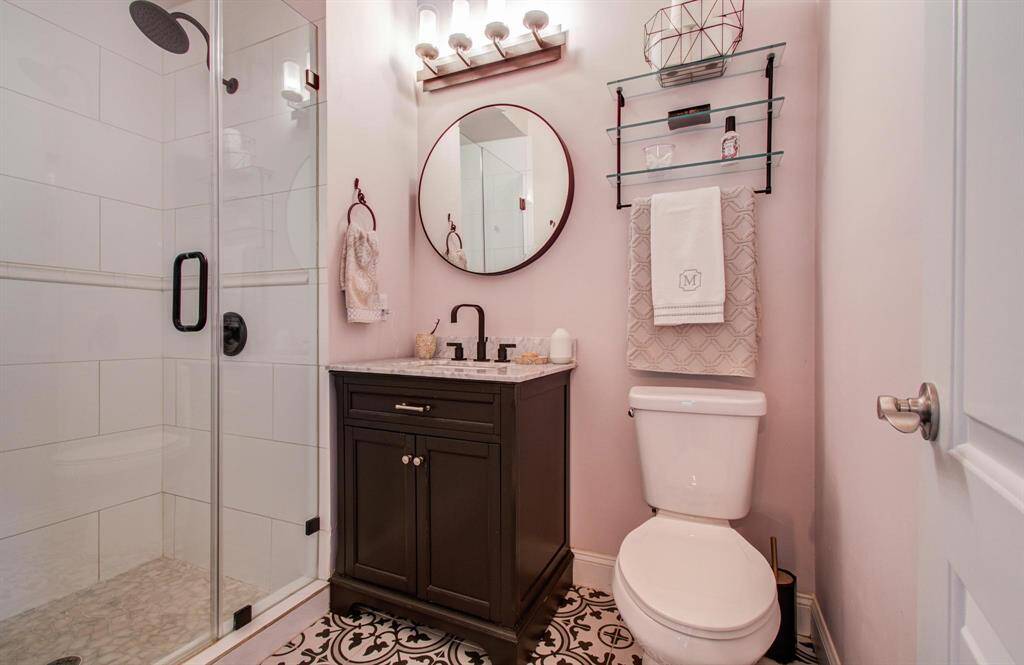 This updated bathroom features a glass-enclosed shower, a sleek vanity with a round mirror, and distinctive tile flooring that reflects the homeowner’s thoughtful eye for design. With convenient shelving and towel storage, the space blends practicality with style in a warm, inviting setting.