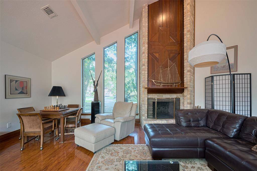 The fabulous main living area is just steps from the front entrance and showcases striking vaulted ceilings, designer wood flooring, and expansive floor-to-ceiling windows that offer picturesque views of the front yard. A charming brick gas-log fireplace serves as the perfect centerpiece, adding warmth and character to the space.