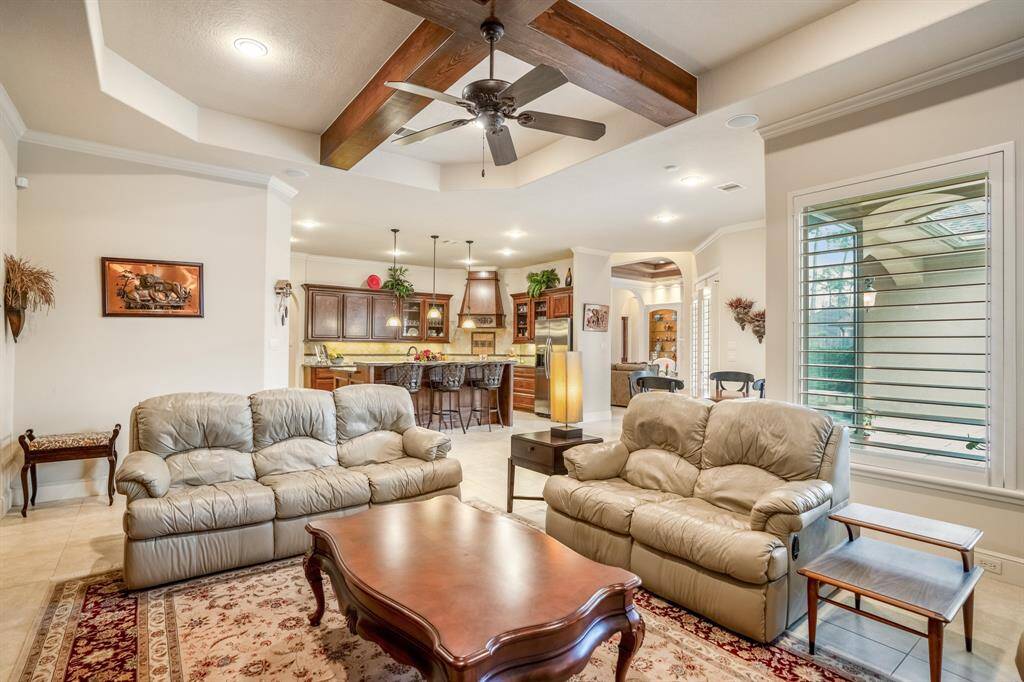 The room flows seamlessly into the open kitchen with rich wooden cabinetry and bar seating, ideal for entertaining. Large windows allow natural light to fill the space, enhancing its airy feel.