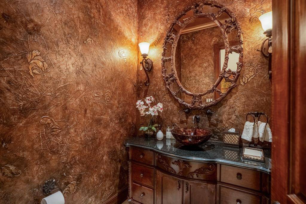 This lavish powder bathroom features rich, textured walls and a luxurious, ornate mirror. The stylish vessel sink with dark granite countertop is complemented by warm lighting and decorative sconces.