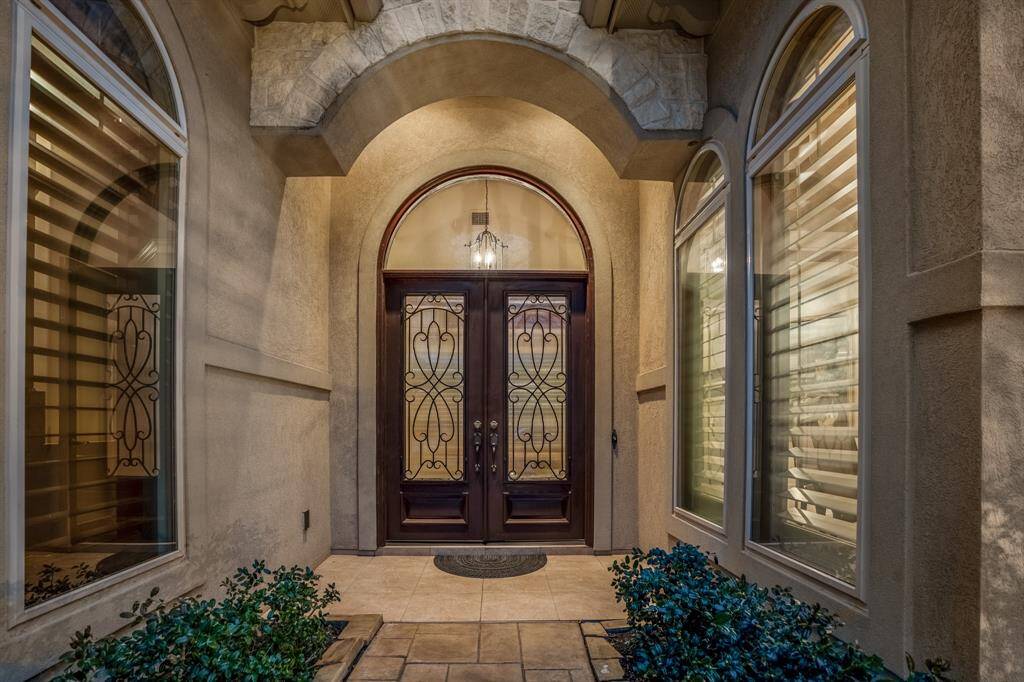The iron-accented elegant double wood doors are flanked by large arched windows. The stone and stucco facade, along with the overhead arch, add a touch of sophistication.