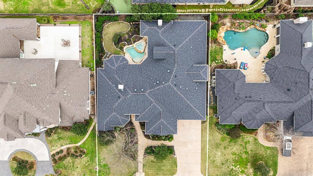 An overhead drone shot captures the entire backyard, highlighting the harmonious layout of the stone-surrounded pool with its cascading waterfalls, integrated hot tub, expansive lanai equipped with an outdoor kitchen, and the surrounding lush green space. Seamless integration of leisure and entertainment areas, illustrating the property's extraordinary features.