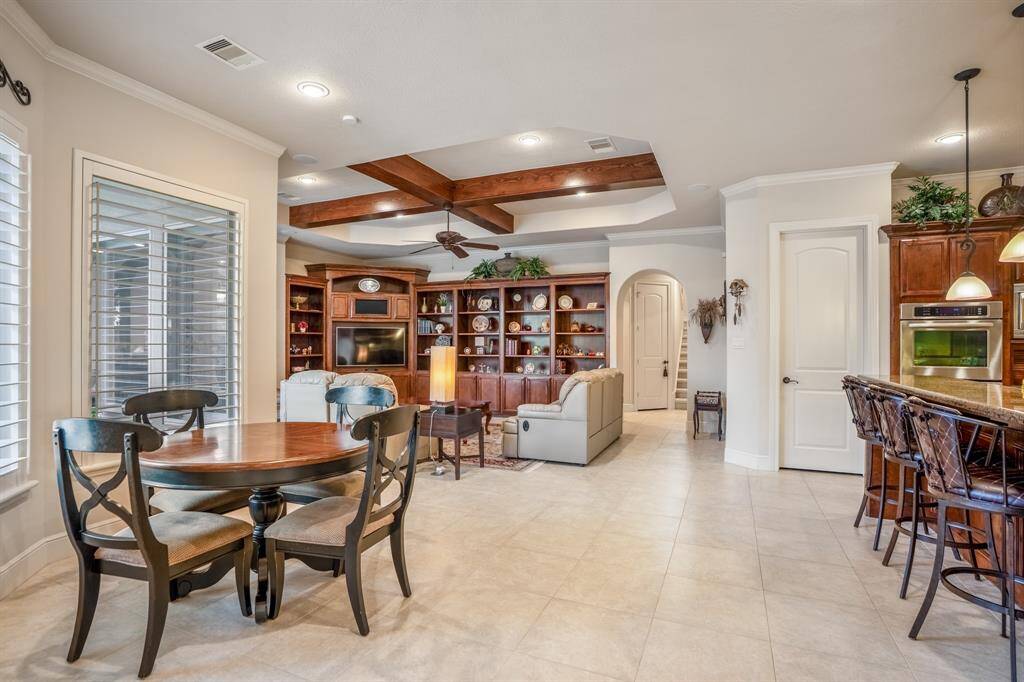 This inviting open-concept 1.5-story layout features a cozy living area with built-in shelving, entertainment center, surround sound throughout, a sun-lit breakfast area, and a large, timeless kitchen. The room is accented by a tray ceiling with wooden beams and large windows for natural light.