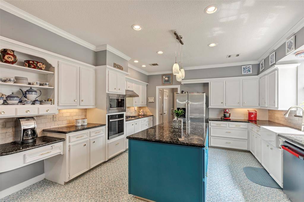 This kitchen features a spacious layout with white cabinetry, modern appliances, and a central island with a dark countertop. The room is well-lit with both natural light and recessed lighting, and has decorative shelving and backsplash for added charm.