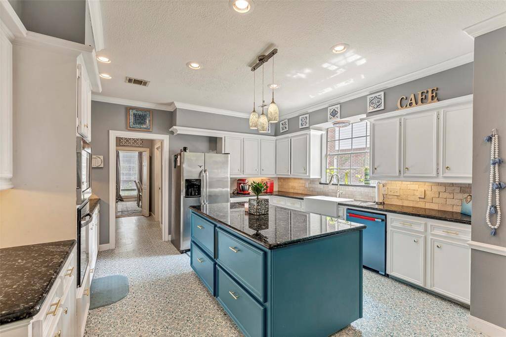 This kitchen features a spacious layout with white cabinetry, modern appliances, and a central island with a dark countertop. The room is well-lit with both natural light and recessed lighting, and has decorative shelving and backsplash for added charm.