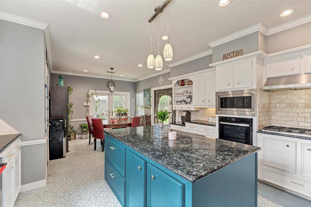 This kitchen features a spacious layout with white cabinetry, modern appliances, and a central island with a dark countertop. The room is well-lit with both natural light and recessed lighting, and has decorative shelving and backsplash for added charm.