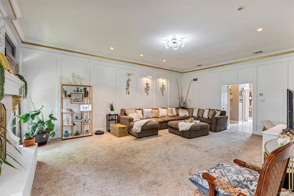 This spacious living room features wooden panels and elegant crown molding. It has a modern chandelier, and ample natural light from large windows, creating a welcoming and stylish atmosphere.