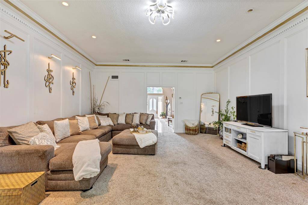 This spacious living room features wooden panels and elegant crown molding. It has a modern chandelier, and ample natural light from large windows, creating a welcoming and stylish atmosphere.