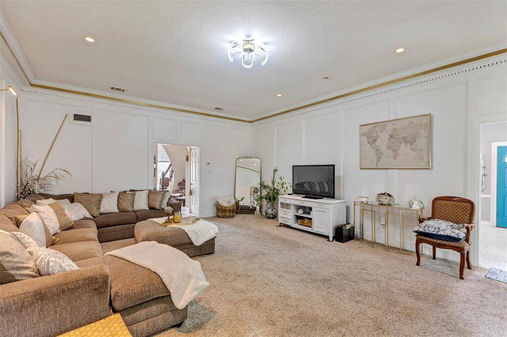 This spacious living room features wooden panels and elegant crown molding. It has a modern chandelier, and ample natural light from large windows, creating a welcoming and stylish atmosphere.