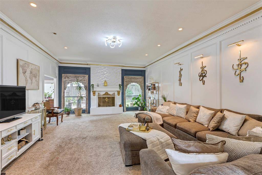 This spacious living room features wooden panels and elegant crown molding. It has a modern chandelier, and ample natural light from large windows, creating a welcoming and stylish atmosphere.