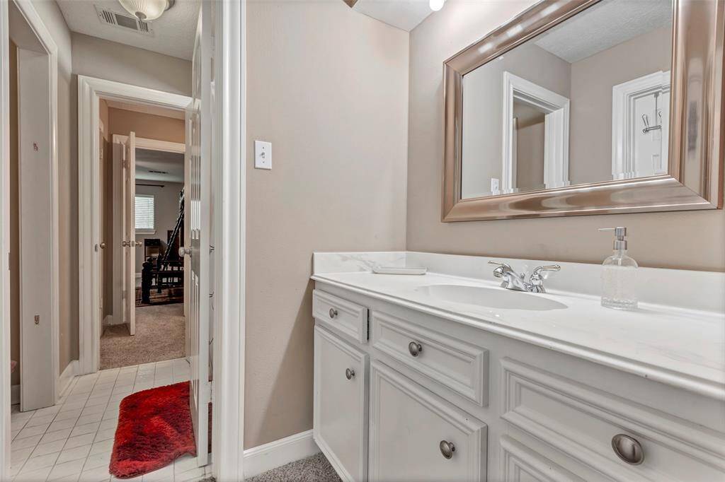 This photo showcases a bright bathroom with a large mirror, a sleek white vanity, and a single sink. The room is complemented by neutral walls and tile flooring, with a glimpse into a carpeted bedroom.