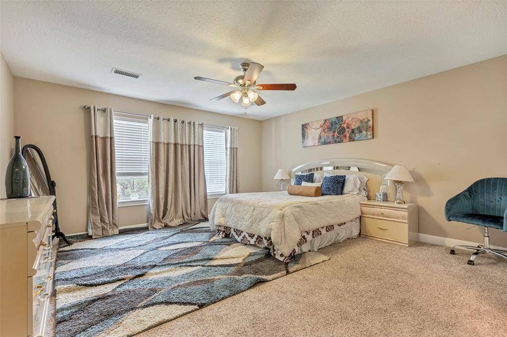 This spacious bedroom features neutral walls, and a large window with elegant drapes. A stylish ceiling fan and a large window which provides plenty of natural light.