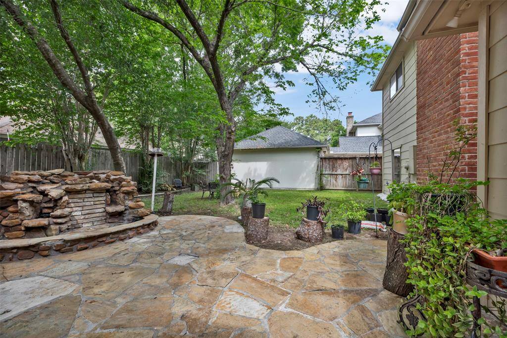 This backyard features a charming stone patio with lush greenery and mature trees. A small rock waterfall adds a tranquil touch, making it an ideal space for relaxation and outdoor entertaining.