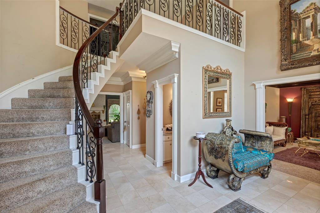 This elegant entryway features a curved staircase with ornate iron railings and plush carpeting. The space is bright and open. Adjacent rooms are visible, offering a glimpse of the home’s luxurious design elements.