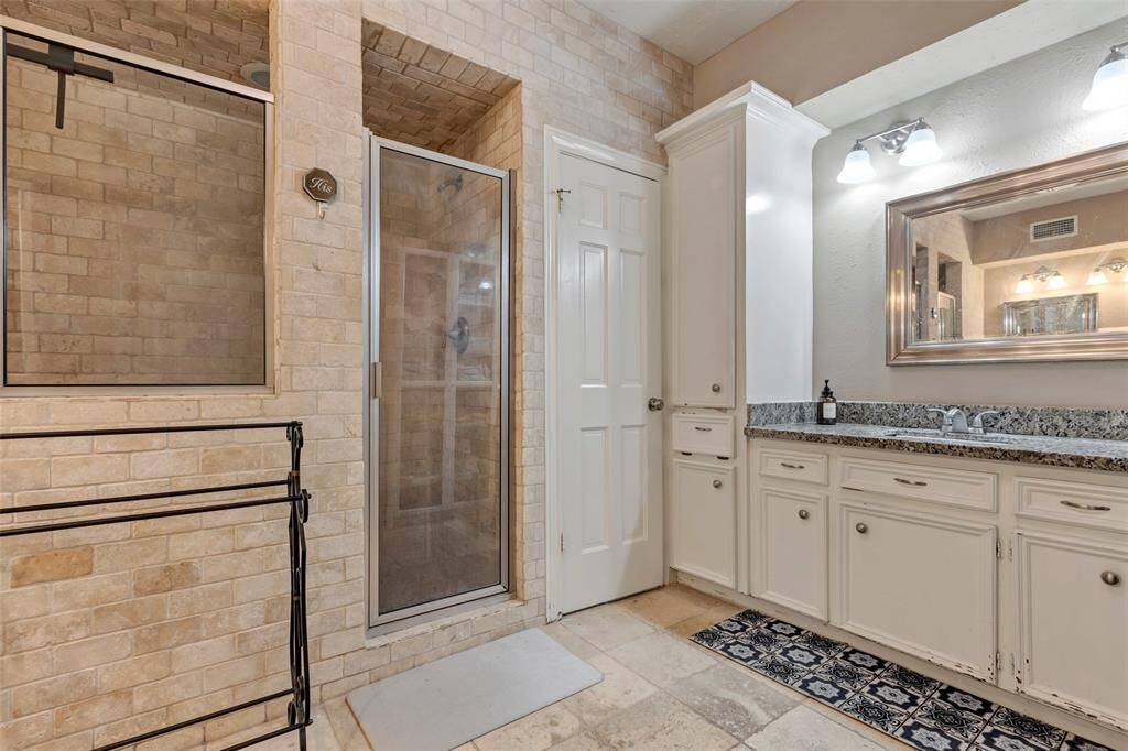 This bathroom features a spacious shower with double glass doors and brick-style tiling. It includes a double vanity with granite countertops and ample cabinet storage. The room is well-lit with modern fixtures and has a neutral palette for a clean, timeless look.