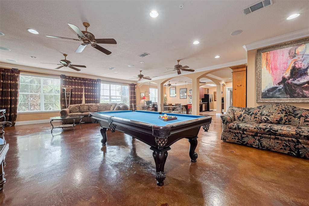 The clubroom invites relaxation and productivity alike with lounge seating, ceiling fans, workstations, and a billiard table, all surrounded by bright windows.