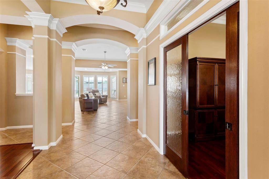 Step inside to a bright, open entryway with elegant arches, crown molding and tile flooring, leading to a spacious living area with abundant natural light. Sliding wood and glass doors open to a home office on the right.