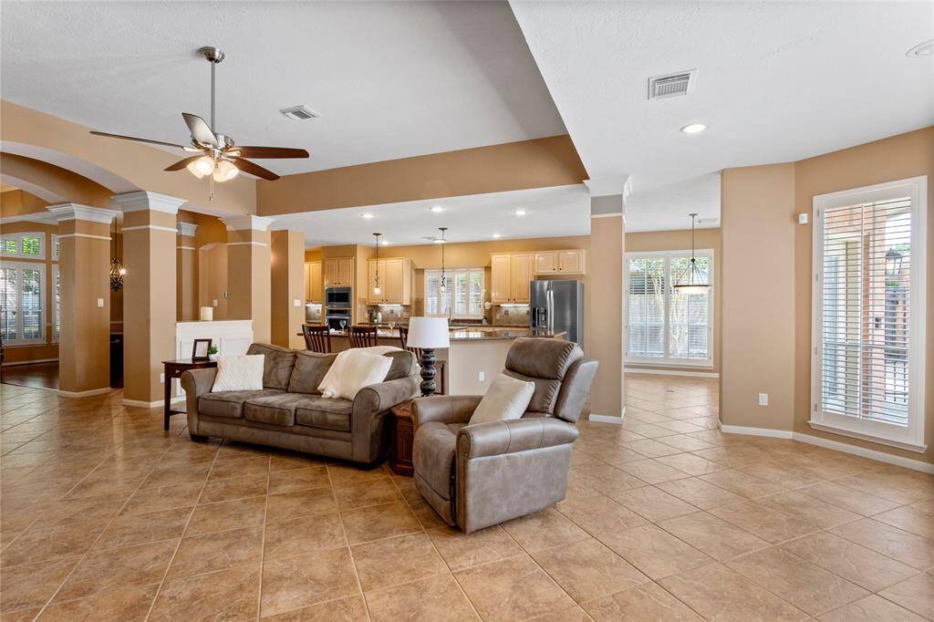 This open-concept living area features a tiled floor, neutral tones, and abundant natural light.  All windows are accented with beautiful wooden plantation shutters. The living room seamlessly connects to the kitchen and dining area. The room is accented by elegant columns and a new ceiling fan, creating a cozy yet stylish atmosphere.