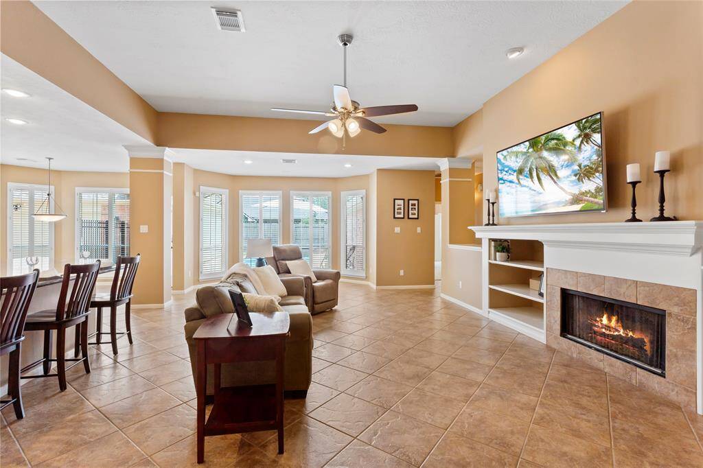 Bright and spacious living area with tile flooring, a cozy fireplace, and a new ceiling fan. It features a modern open layout connecting to the kitchen with bar seating and kitchen, perfect for entertaining. Large windows provide ample natural light, creating an inviting atmosphere and views to the backyard pool.