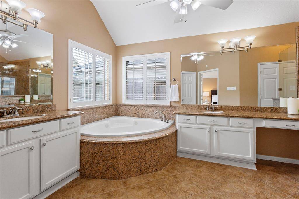 The Primary Ensuite Bathroom features a spacious layout with a luxurious corner jetted bathtub surrounded by granite countertops and solid wood cabinetry. Includes dual sinks with ample countertop space and vanity space. Large mirrors and windows with plantation shutters provide plenty of natural light, complemented by elegant lighting fixtures.