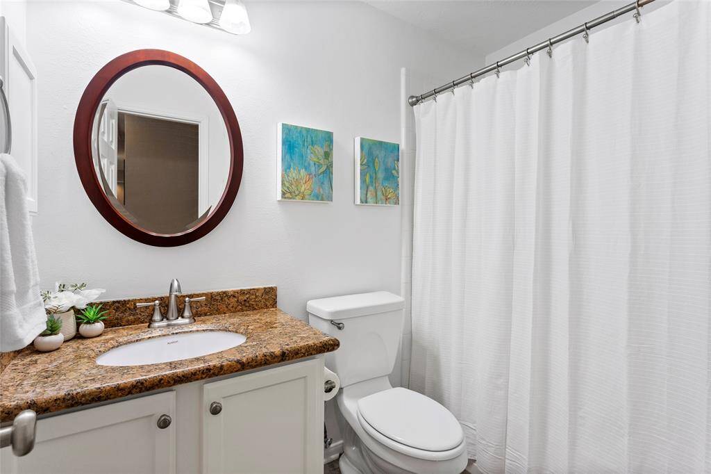 This full bathroom features a clean, modern design with a granite countertop and white cabinetry. It includes a round mirror and stylish lighting.